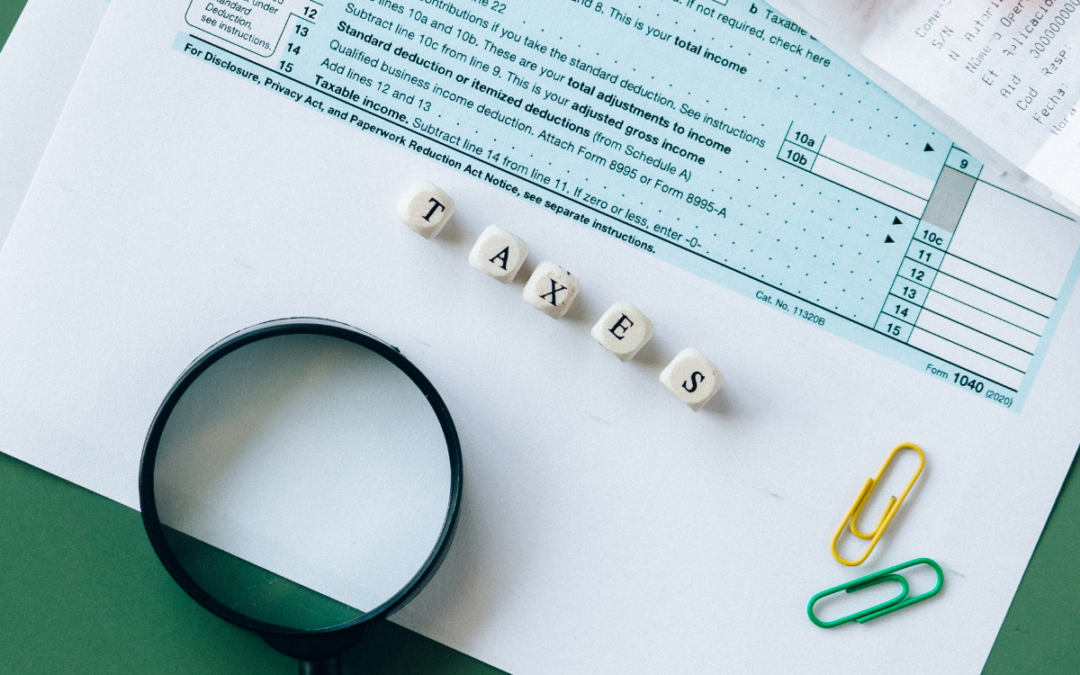 Close-up of tax forms, a magnifying glass, and paper clips on a table. White cubes spell out the word "TAXES